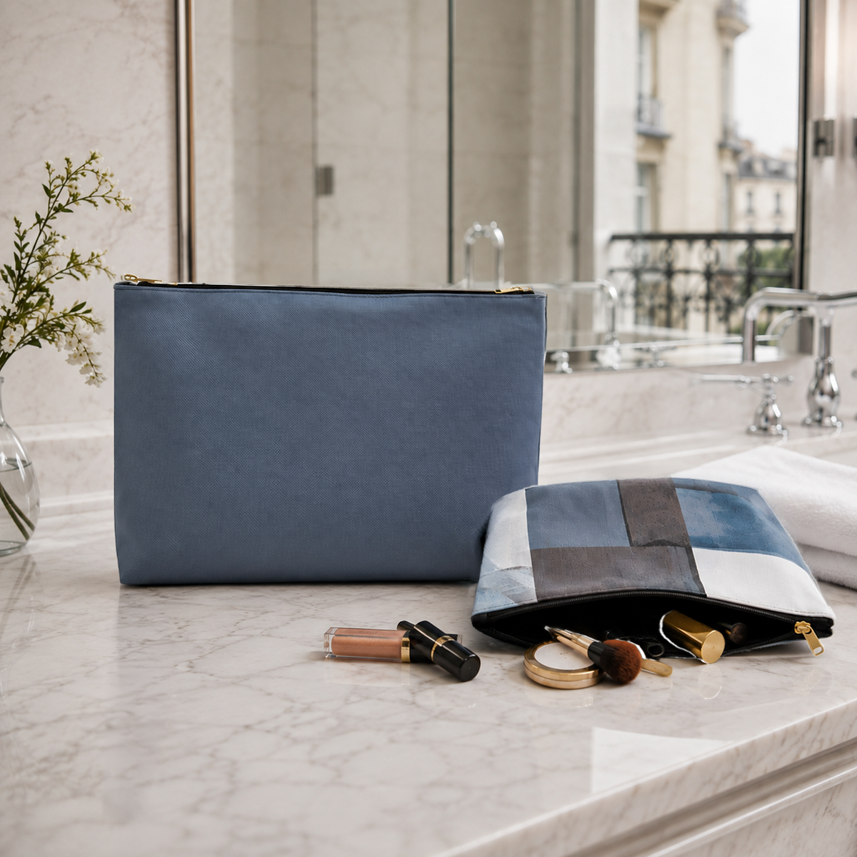 A Slate Blue zippered cosmetic pouch sits on a marble bathroom counter beside a Slate and Blue color block pouch, with cosmetics and a folded towel nearby. A mirror, sink, and flowers are visible in the background.