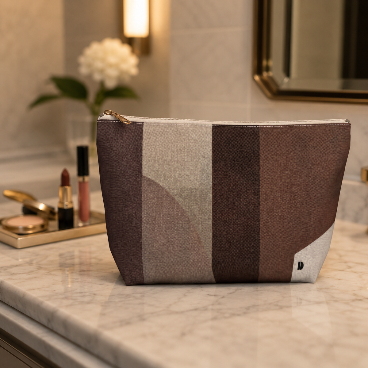 A Study in Brown T-bottom Accessory Pouch featuring an earth-toned geometric pattern sitting on a marble bathroom vanity next to makeup products.