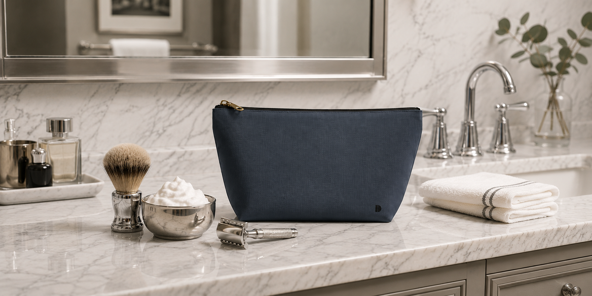 A Slate and Blue zippered toiletry pouch sits on a marble bathroom counter, surrounded by shaving accessories, a folded white towel, and a chrome faucet in the background.