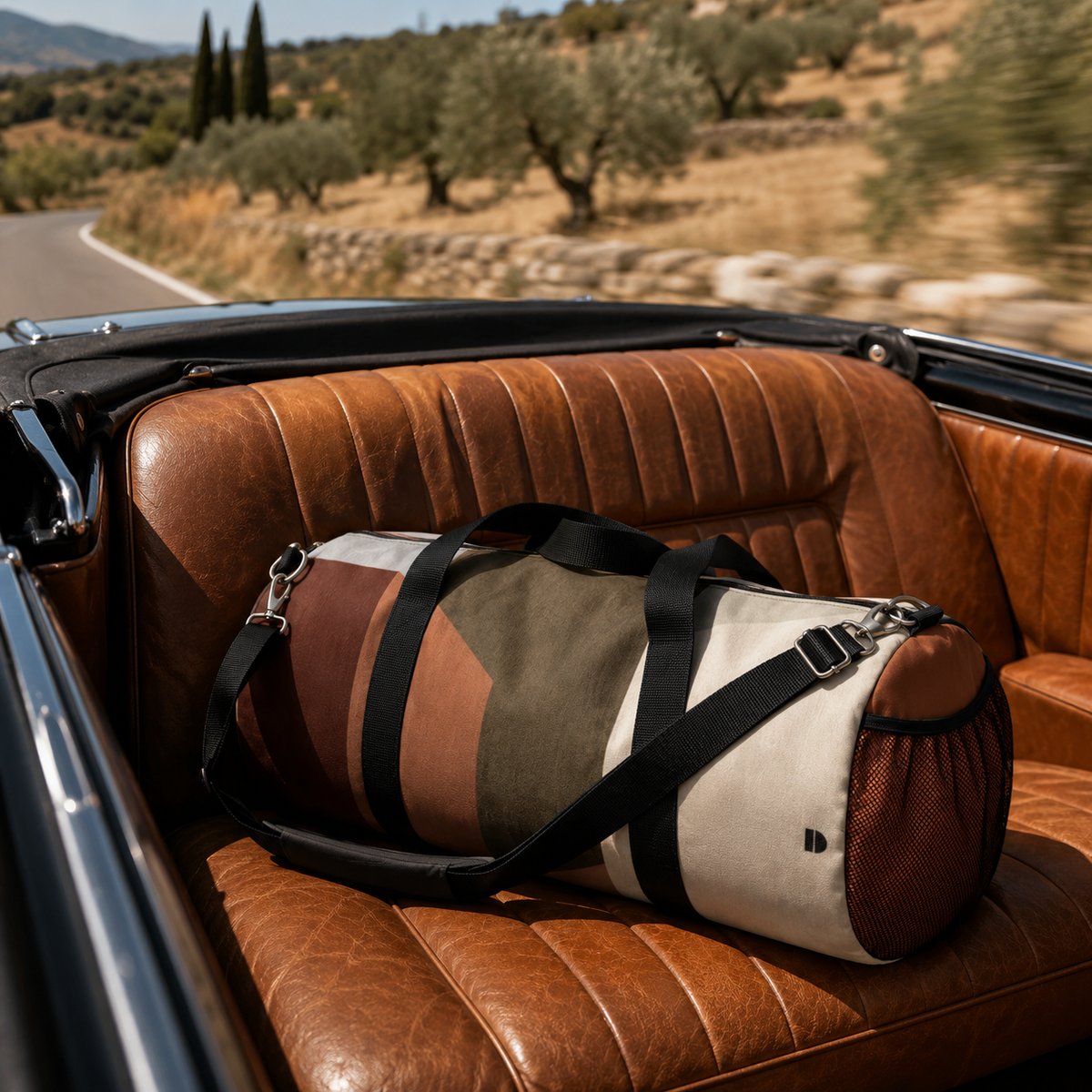A Study in Brown Duffel with a color block design in brown, beige, and olive green, featuring black straps, resting on the brown leather backseat of a convertible car on a road with a blurred Mediterranean landscape background.