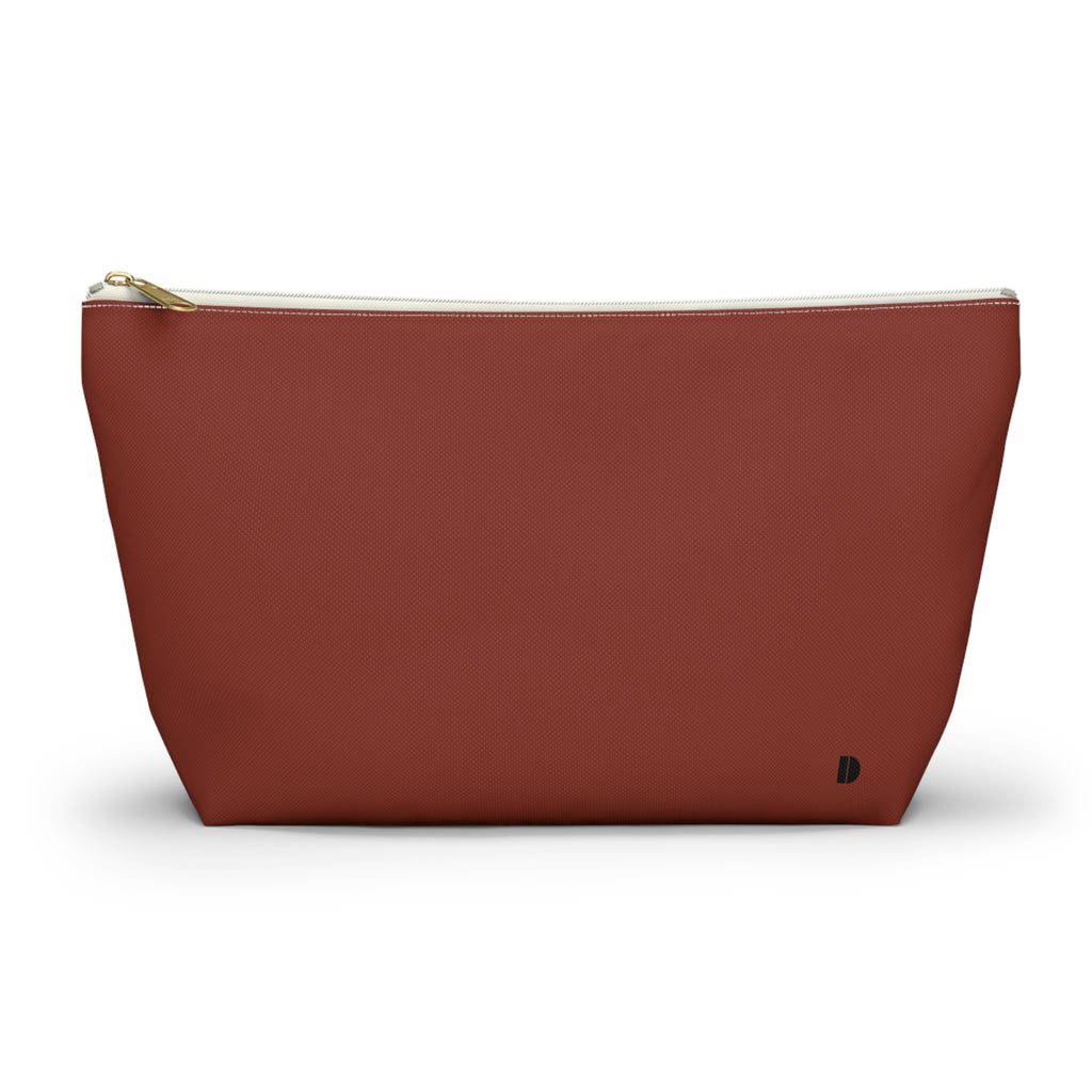A terracotta red canvas cosmetic pouch with a gold zipper and a small black monogram logo on the bottom right, set against a plain white background.