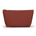 A terracotta red canvas cosmetic pouch with a gold zipper and a small black monogram logo on the bottom right, set against a plain white background.