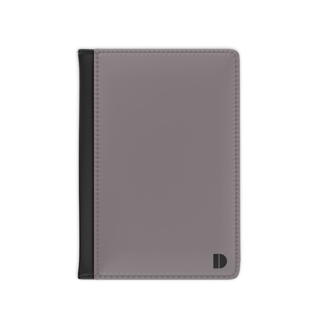 A minimalist passport cover or document holder in a muted grayi with a black spine band and a small black monogram logo in the bottom right corner, shown on a plain white background.