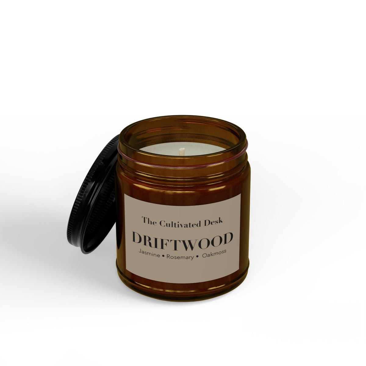 Woody scented candle in an open 4oz amber glass jar showing the white wax and single wick inside, with the black metal lid leaning against the side, featuring a minimalist warm tan label with black serif text reading "The Cultivated Desk," "DRIFTWOOD," and scent notes "Jasmine • Rosemary • Oakmoss." Scented candle titled 'Driftwood', available in 9oz and 4oz sizes.