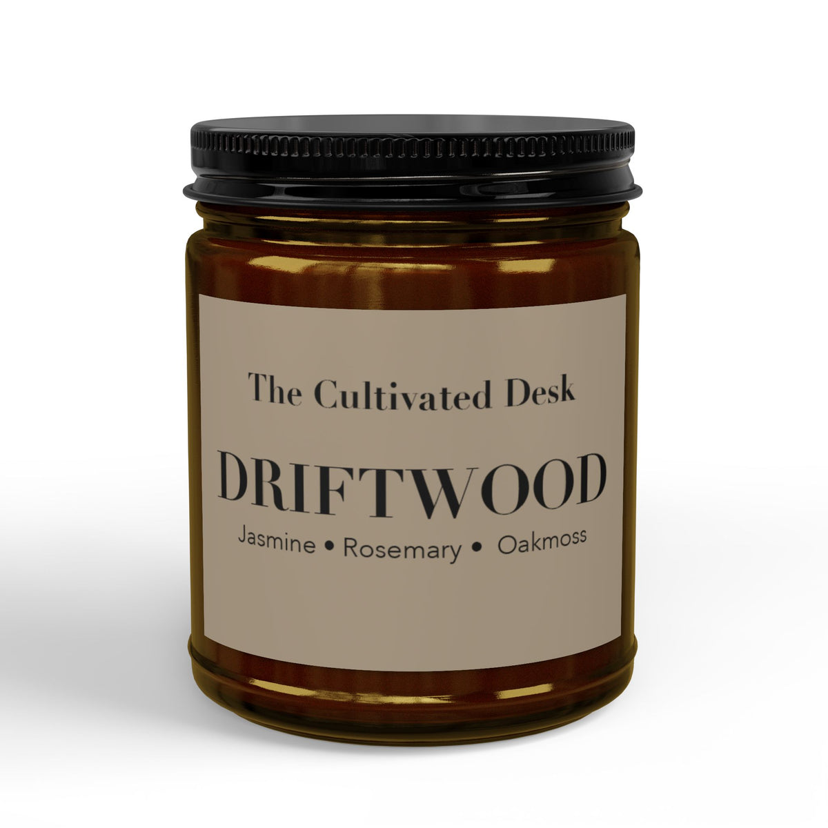 Woody scented candle in a 9oz amber glass jar featuring a minimalist warm tan label with the text "The Cultivated Desk," "DRIFTWOOD," and scent notes "Jasmine • Rosemary • Oakmoss" in black serif typography, sealed with a ribbed black metal lid. Scented candle titled 'Driftwood', available in 9oz and 4oz sizes.