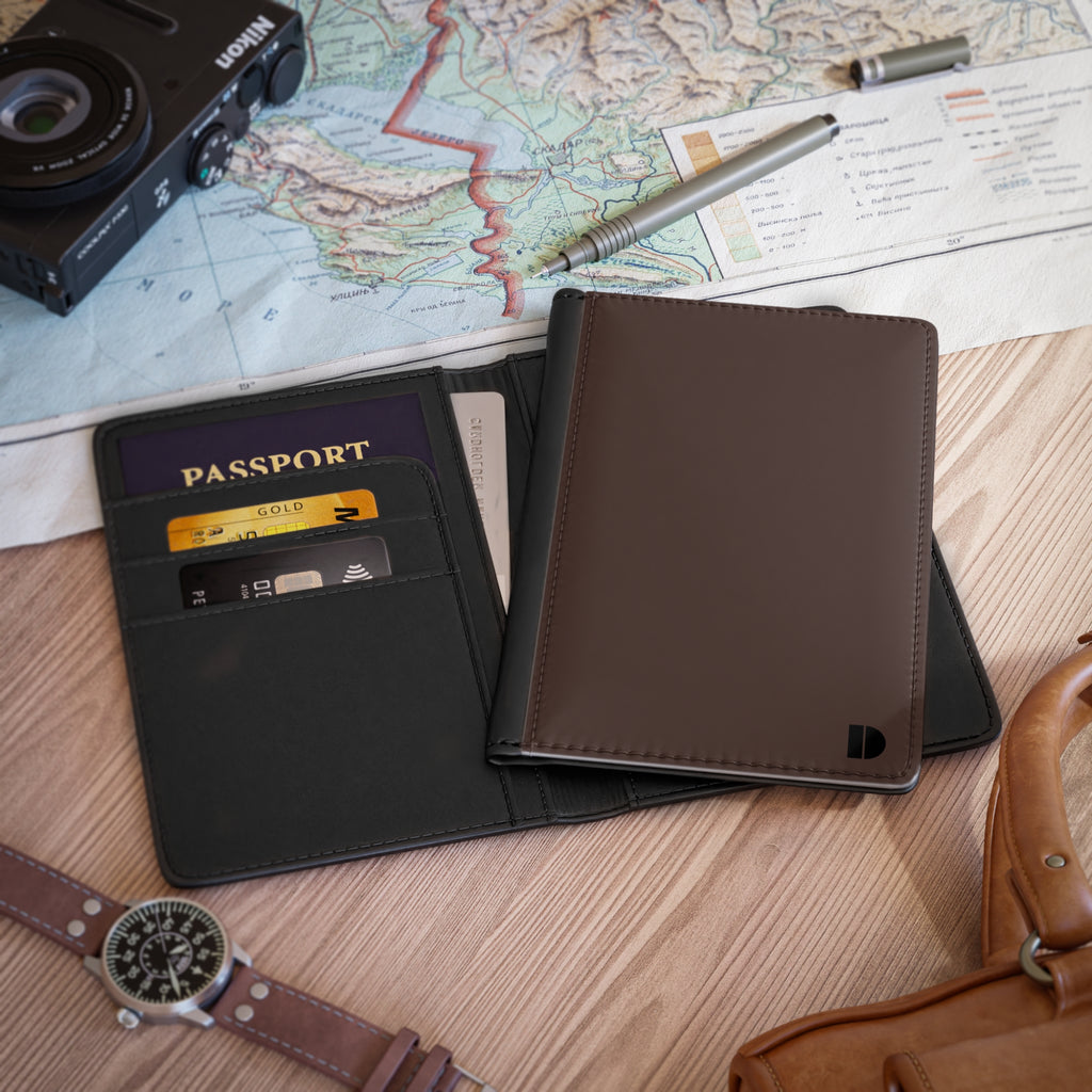 A dark brown faux leather passport holder wallet shown open on a wooden tabletop, with a passport, cards, pen, camera, map, and watch around it as lifestyle props.