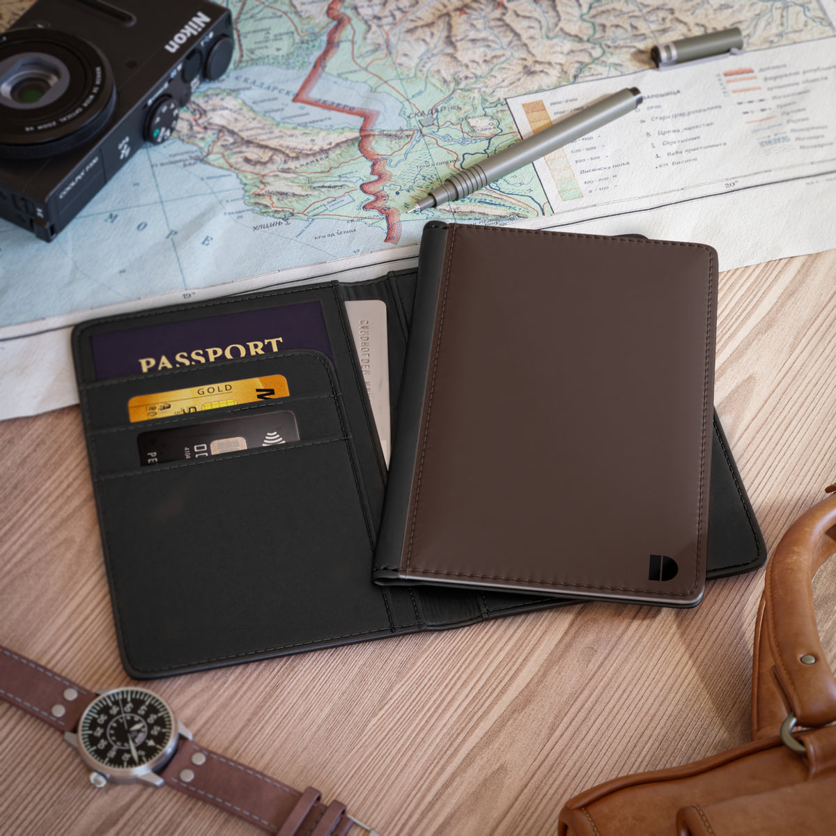 A dark brown faux leather passport holder wallet shown open on a wooden tabletop, with a passport, cards, pen, camera, map, and watch around it as lifestyle props.