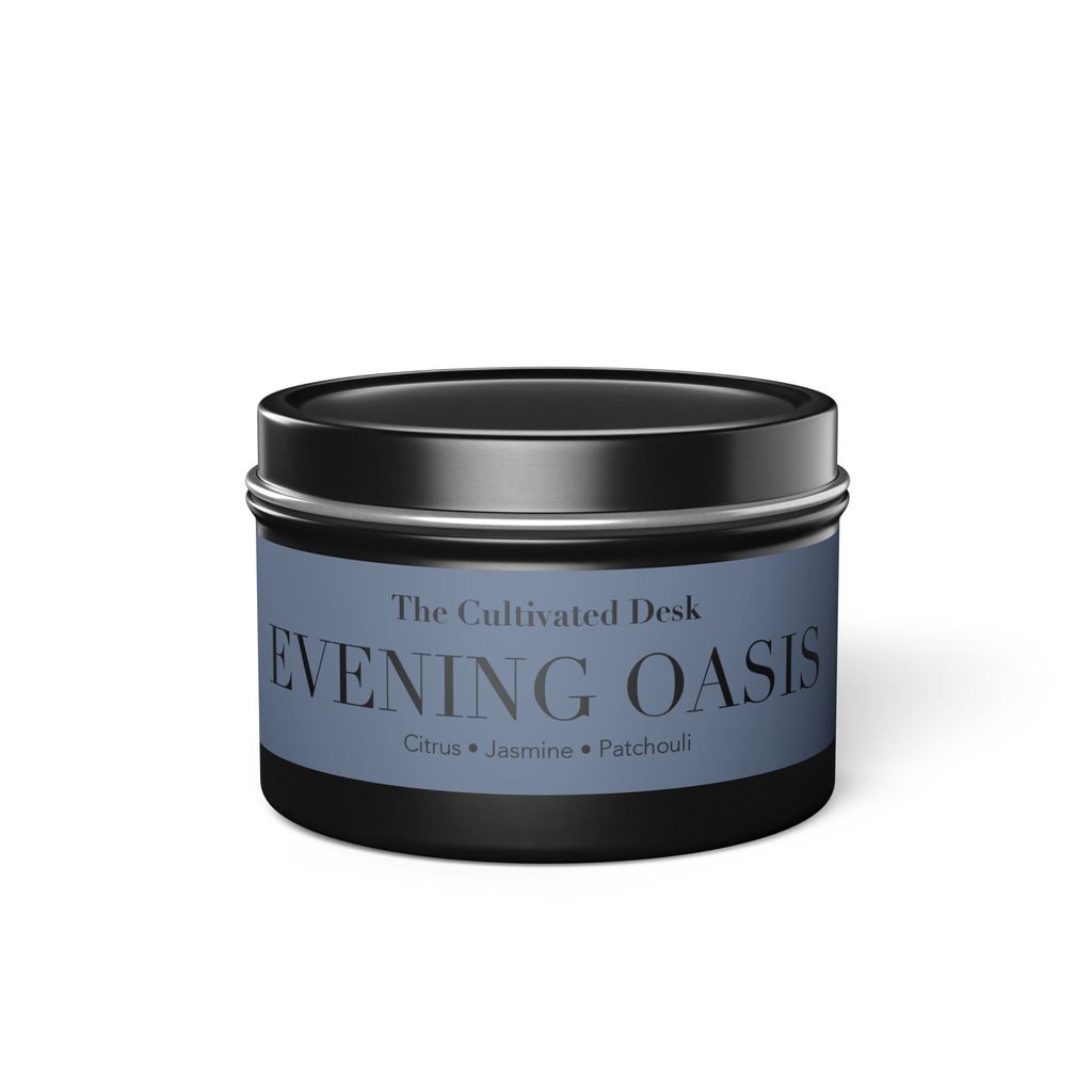 A round metal tin candle with a black lid and base, featuring a dusty blue label that reads 'The Cultivated Desk EVENING OASIS' with citrus, jasmine, and patchouli notes.