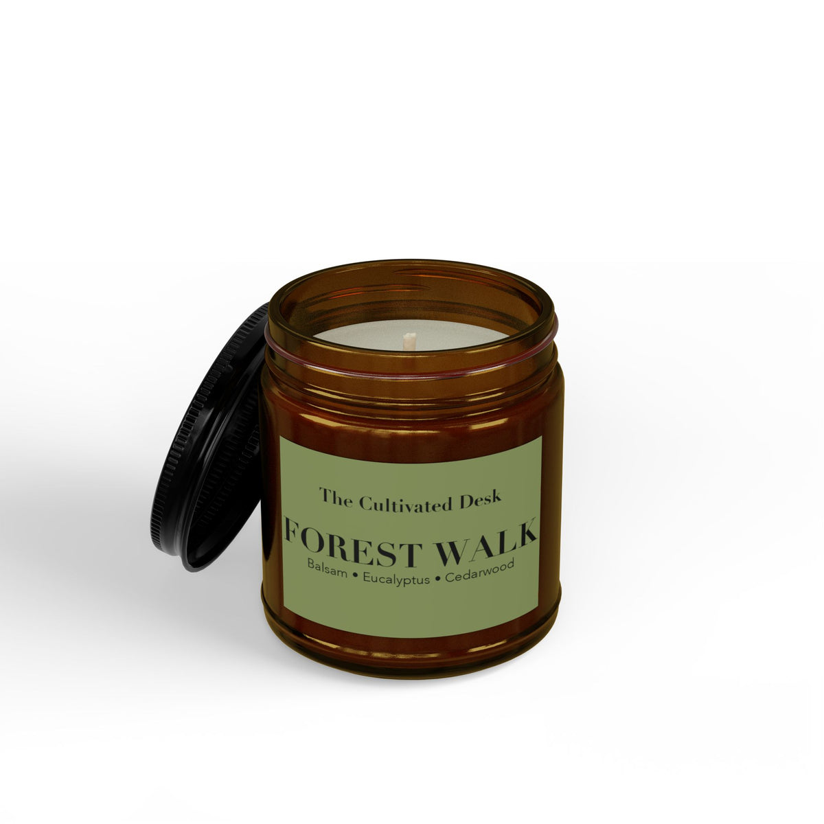 Earthy scented candle in an open 4oz amber glass jar showing the white wax and single wick inside, with the black metal lid leaning against the side, featuring a minimalist sage green label with black serif text reading "The Cultivated Desk," "FOREST WALK," and scent notes "Balsam • Eucalyptus • Cedarwood." Scented candle titled 'Forest Walk', available in 9oz and 4oz sizes.