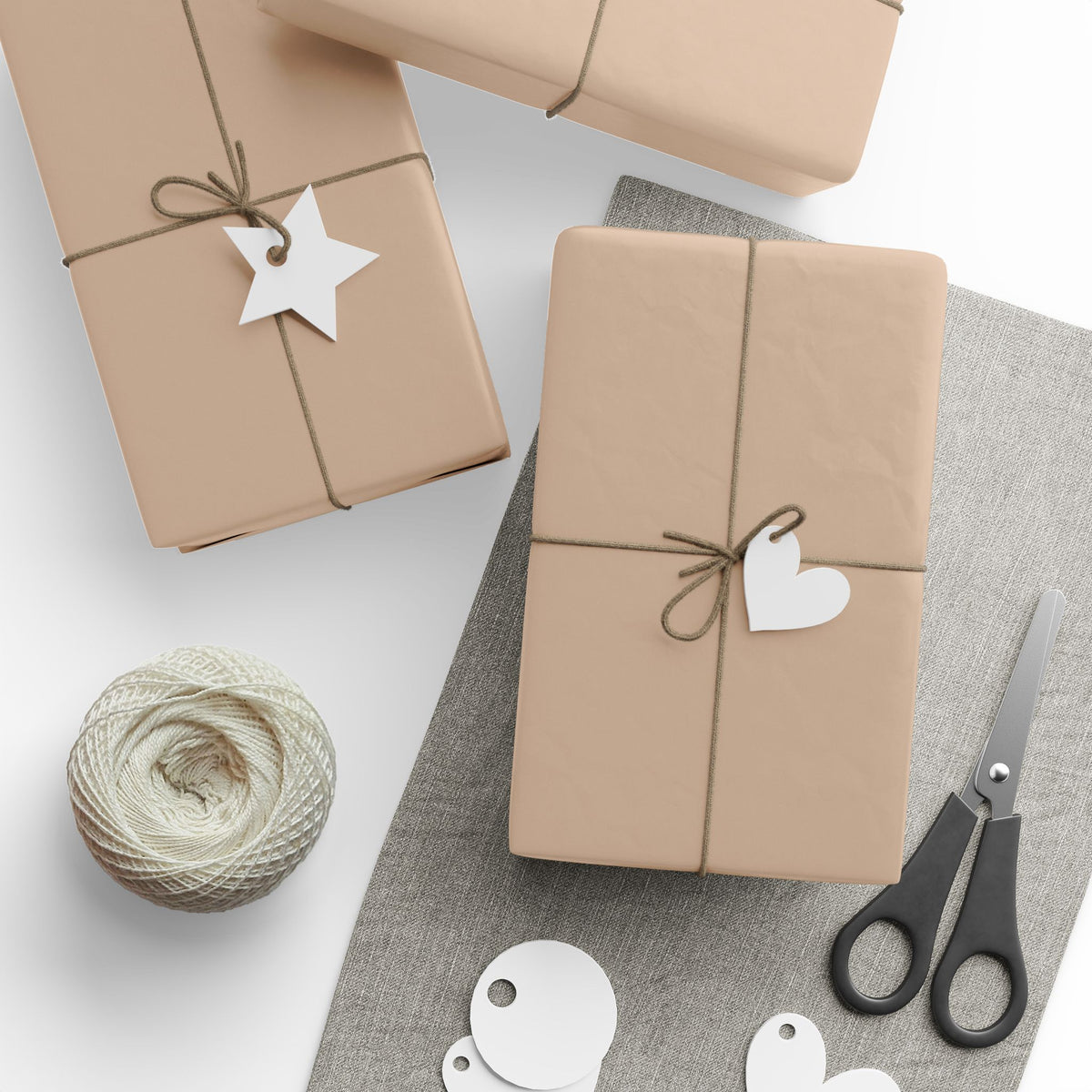 Three gift boxes wrapped in tan wrapping paper with twine bows and white paper tags, arranged on a white and gray surface with a spool of twine and scissors nearby.