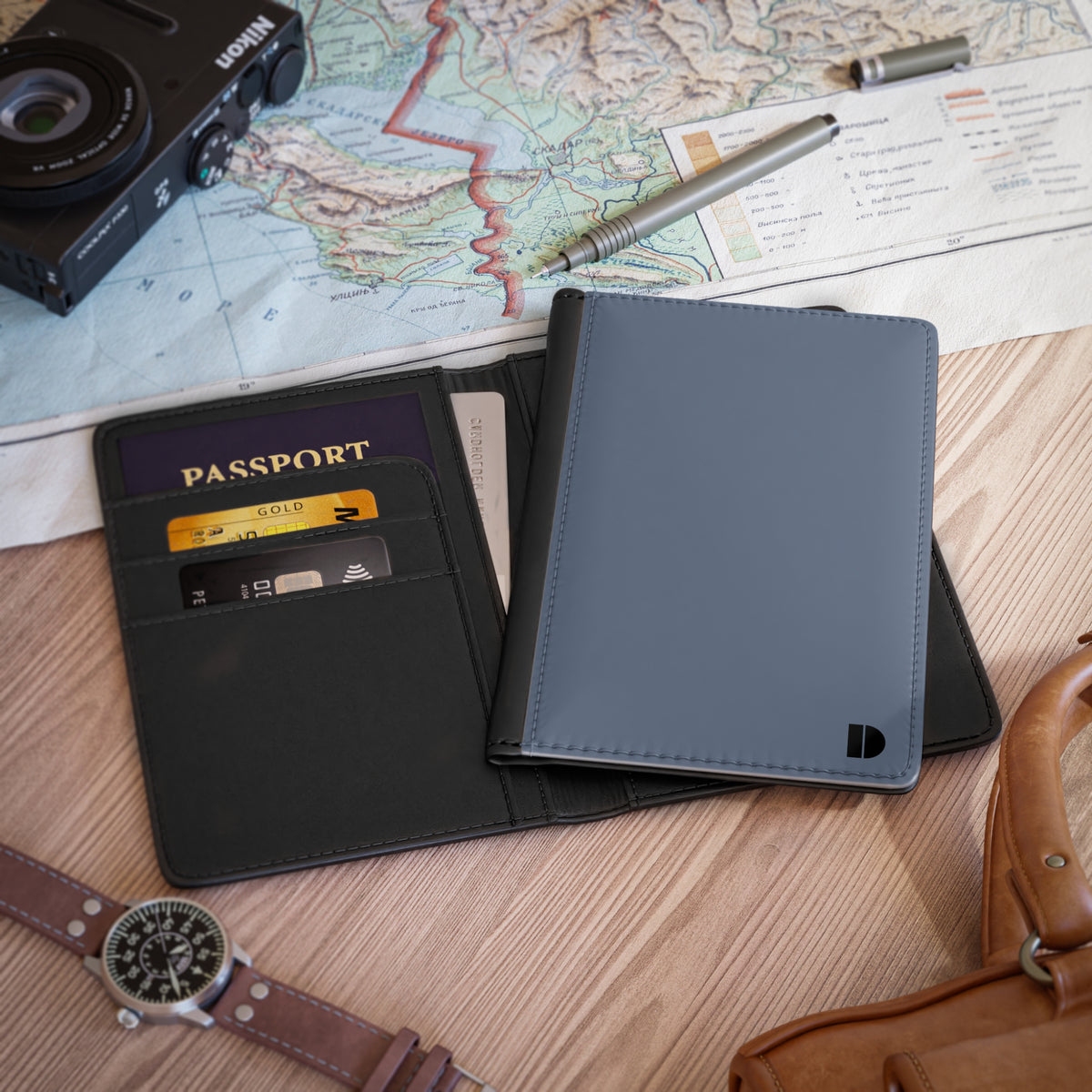 A blue faux leather passport holder wallet shown open on a wooden tabletop with travel props, including a map, camera, pen, watch, and leather bag. The wallet contains cards and a passport.