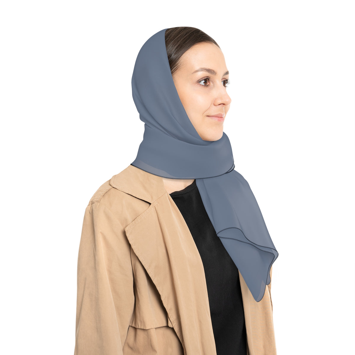 A woman in profile wearing a slate blue wrap as a head scarf and a beige trench coat over a black top, photographed against a solid white background.