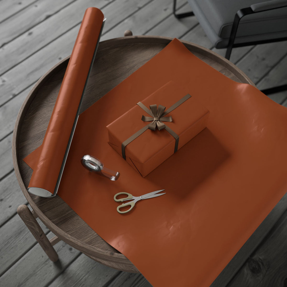 A roll of rust-orange wrapping paper and a matching wrapped gift box with a bow, arranged on a round wooden tray with scissors on a gray deck.
