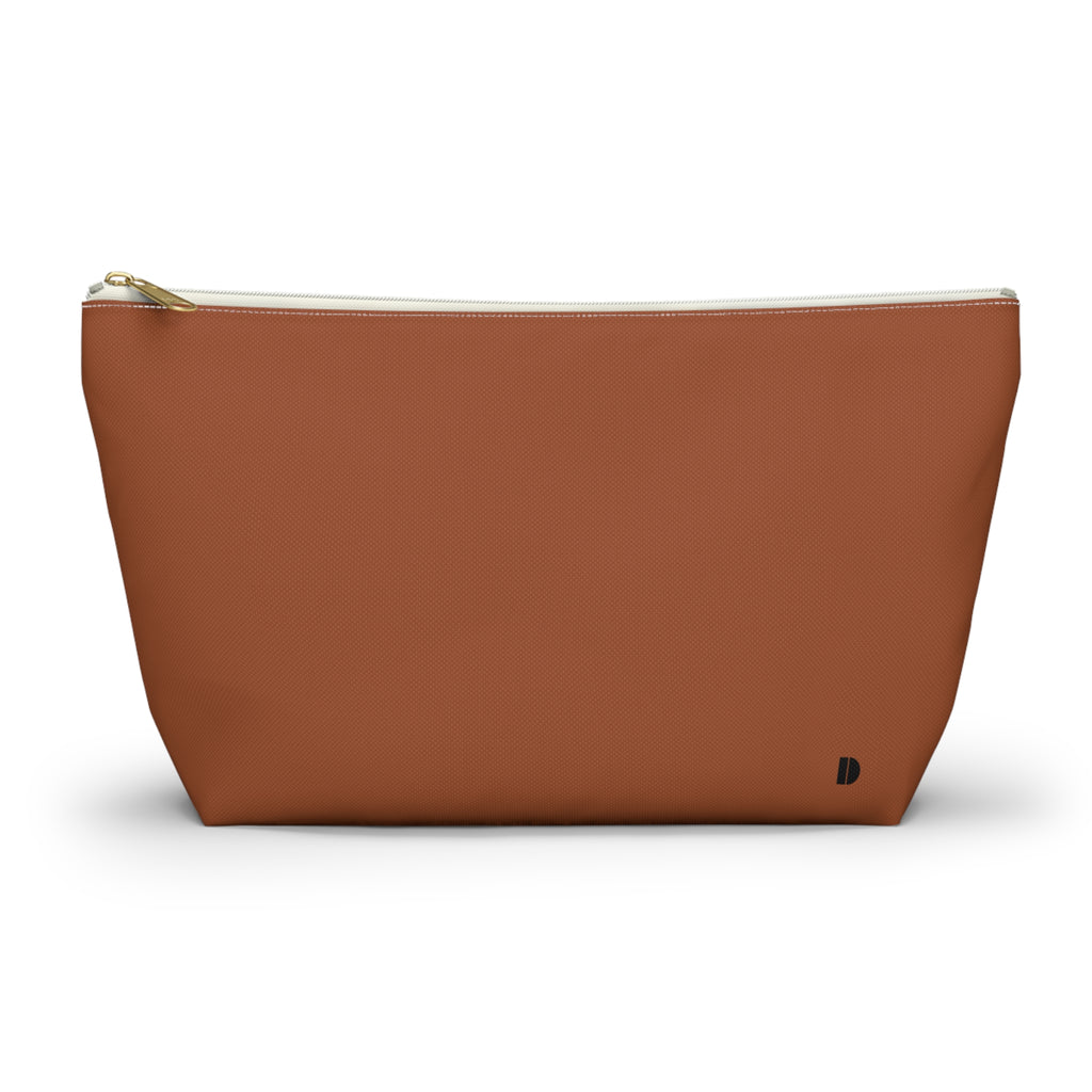 A rust-colored cosmetic pouch with a gold zipper and a small black monogram logo on the bottom right, set against a plain white background.