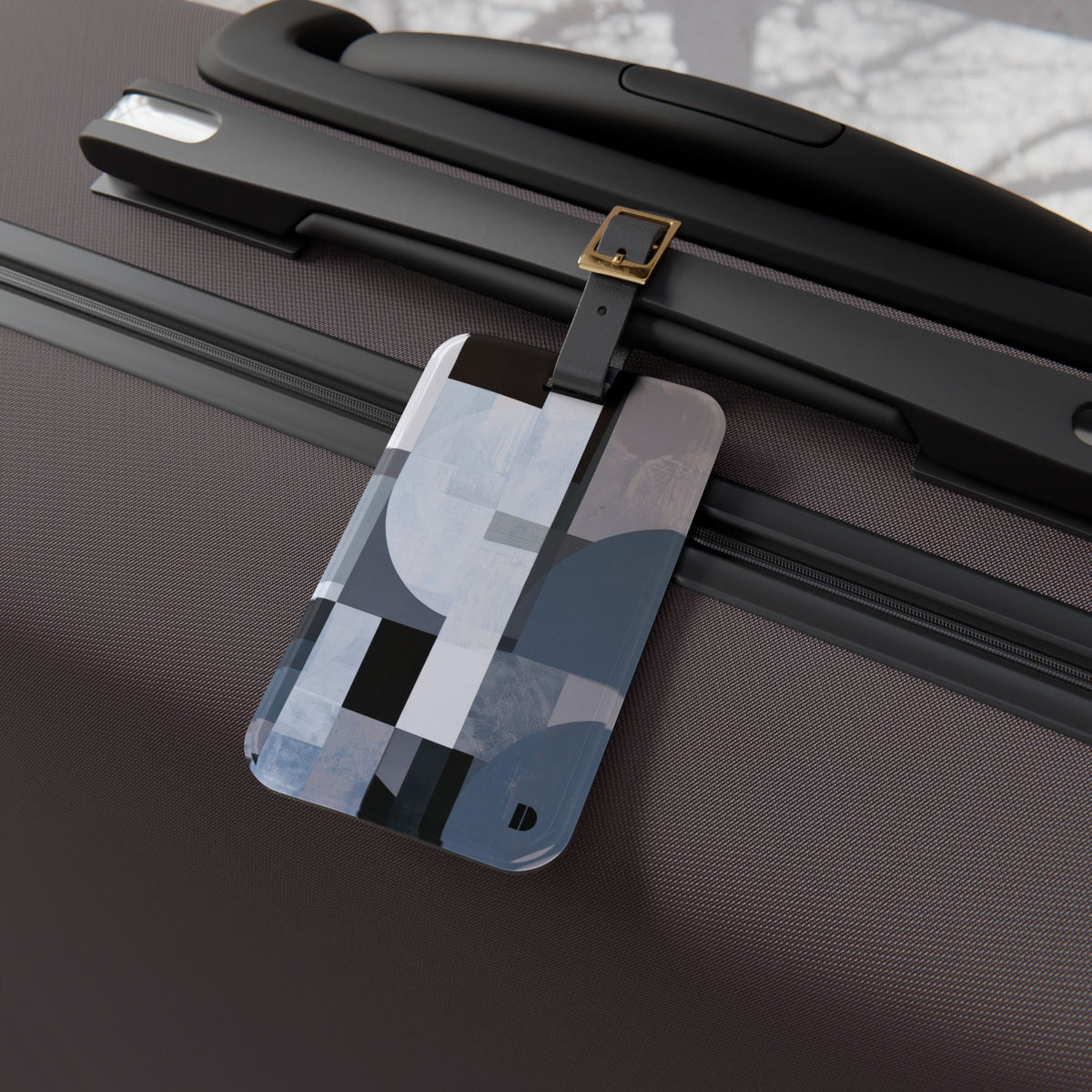 A rectangular luggage tag with an abstract geometric pattern in shades of blue, gray, white, and black, attached to a black strap on a dark brown suitcase.