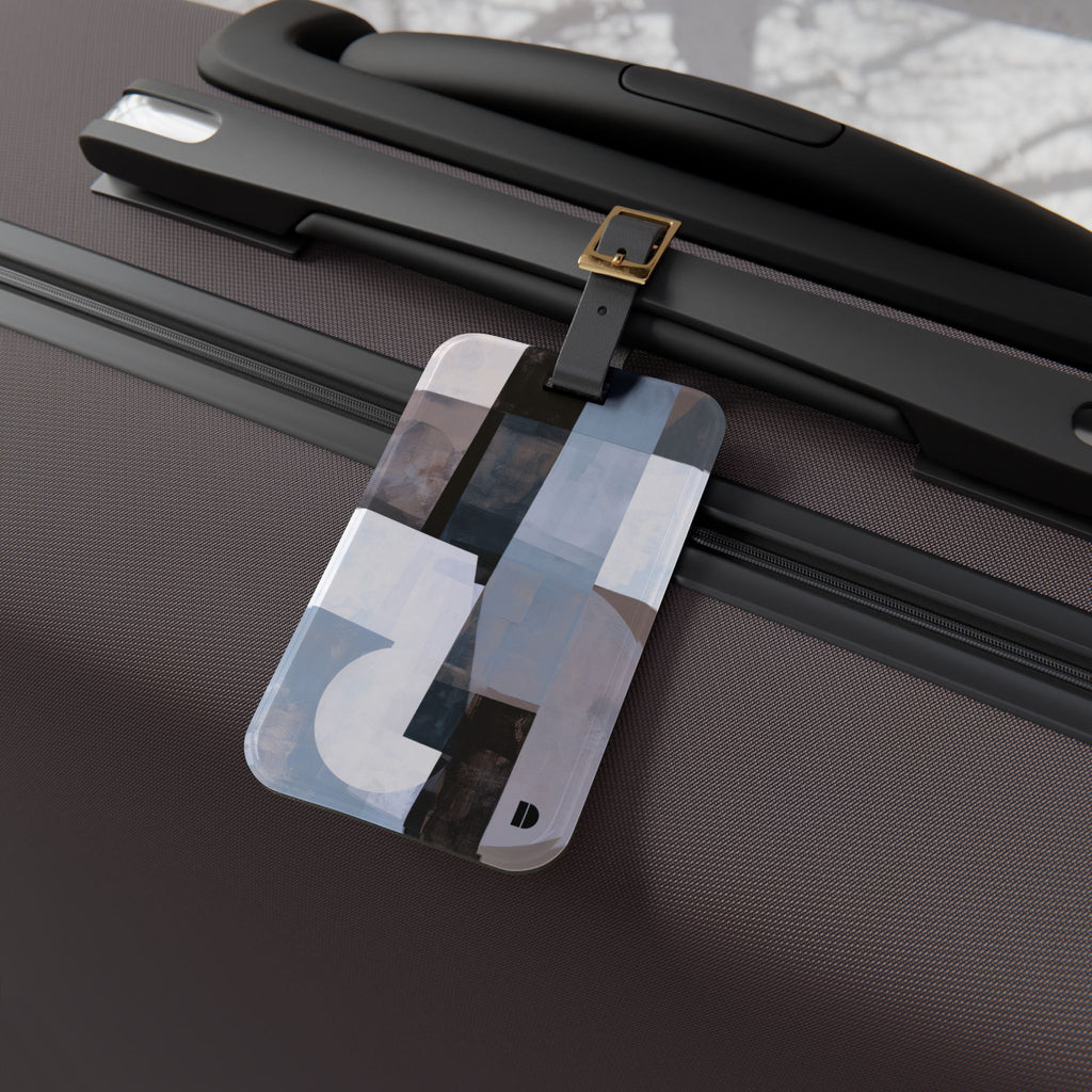 A rectangular luggage tag with an abstract geometric pattern in shades of gray, white, and black, attached to a black strap with a gold buckle on a dark brown textured suitcase.