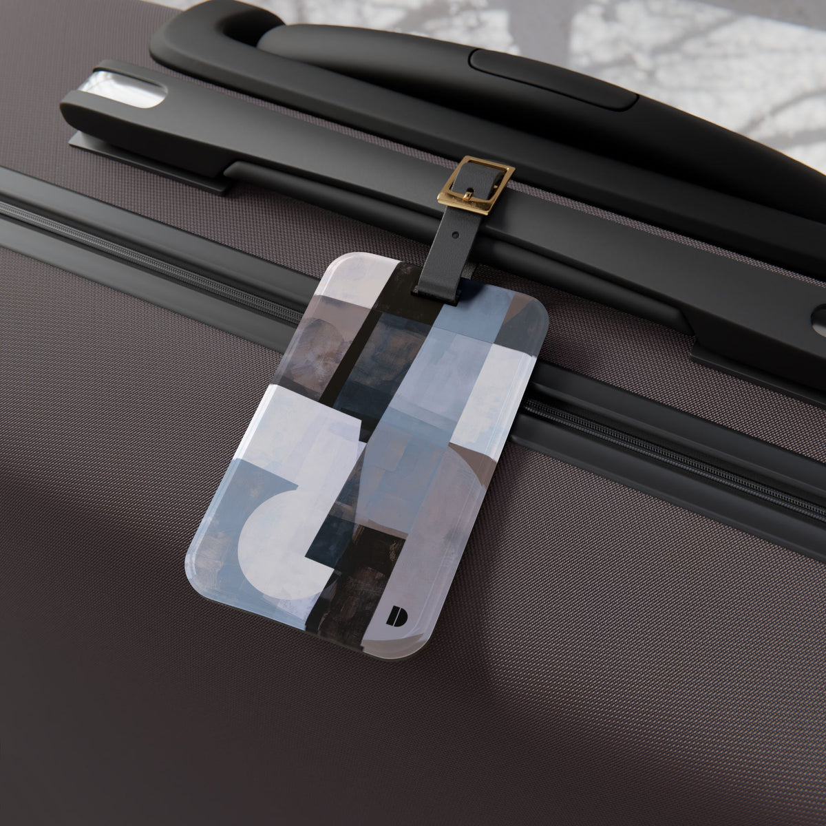 A rectangular luggage tag with an abstract geometric pattern in shades of gray, white, and black, attached to a black strap with a gold buckle on a dark brown textured suitcase.