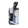A rectangular luggage tag with rounded corners, featuring an abstract geometric pattern in shades of blue, gray, black, and white. It has a black leather strap attached at the top with a gold-tone metal buckle.