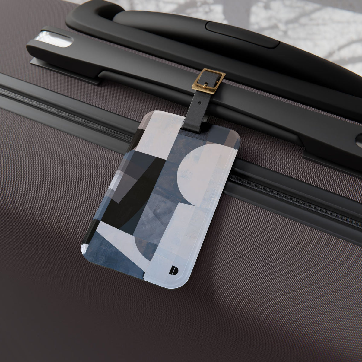 A rectangular luggage tag with an abstract geometric pattern in black, white, and blue-gray, attached to a black strap on a dark brown textured suitcase.