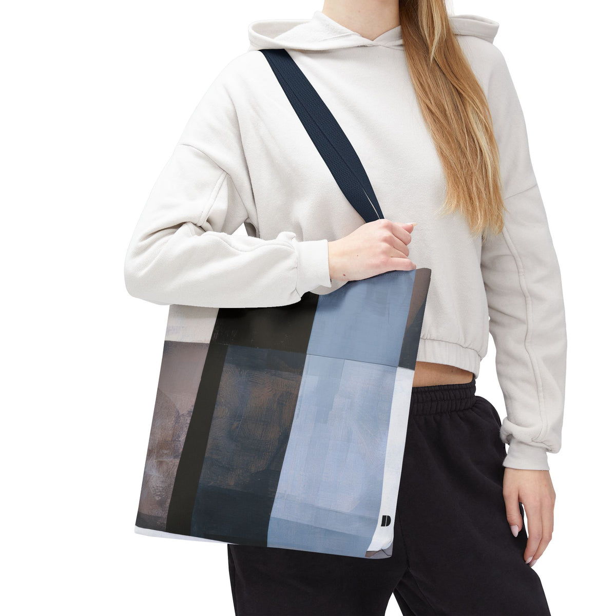 A woman wearing a white hoodie, and black sweatpants carries a large canvas tote bag with a geometric patchwork pattern in shades of blue, black, and gray. The photo is shot against a solid white background.