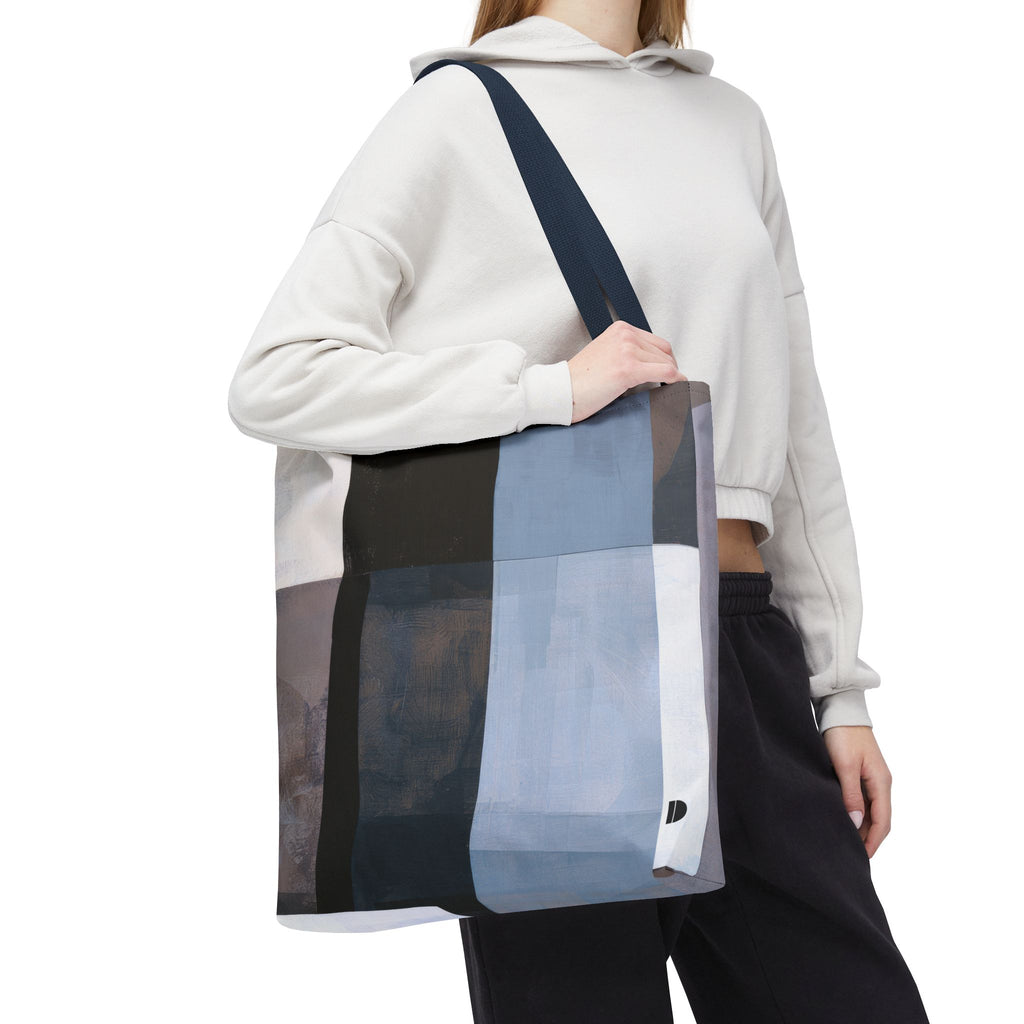 A person wearing a white cropped hoodie and black pants carries a large patchwork tote bag with a dark navy shoulder strap. The tote bag features large rectangular panels in black, gray, light blue, and white.