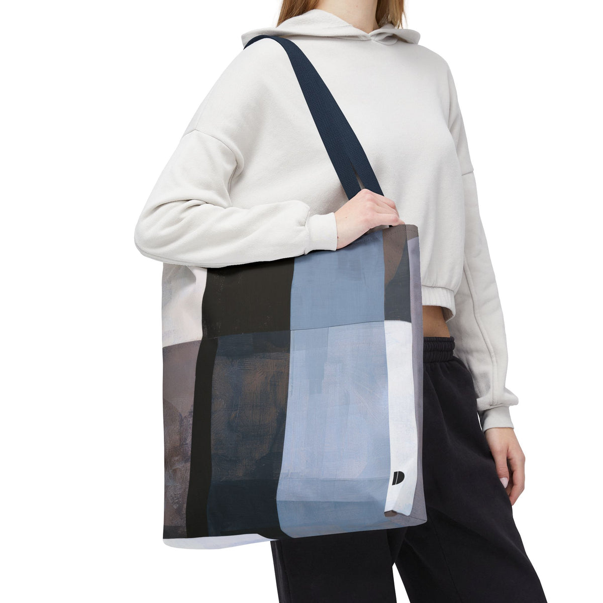 A person wearing a white cropped hoodie and black pants carries a large patchwork tote bag with a dark navy shoulder strap. The tote bag features large rectangular panels in black, gray, light blue, and white.