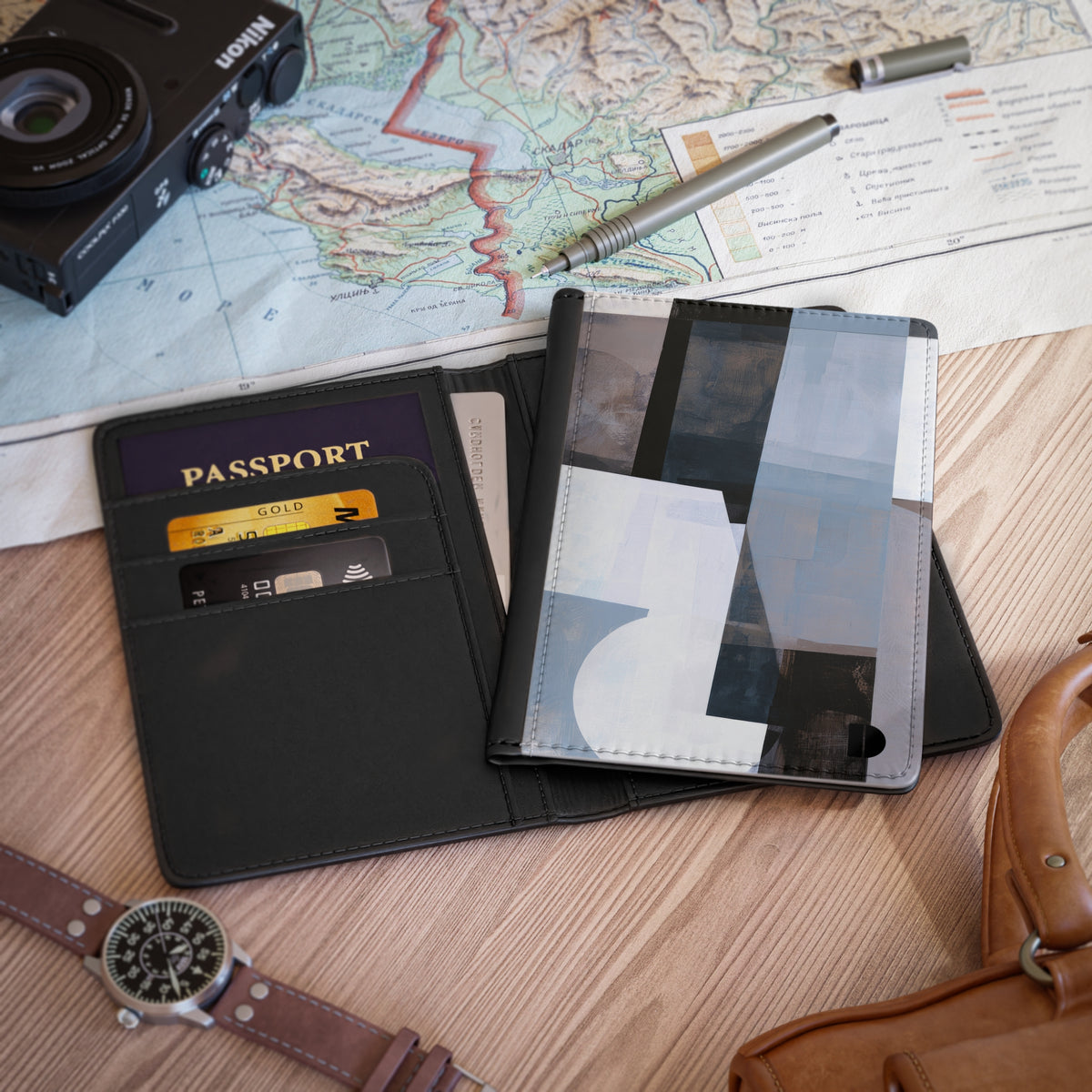 A faux leather passport holder and card case shown open on a wooden surface, with a passport, cards, a map, camera, pen, watch, and leather bag nearby as styled props.