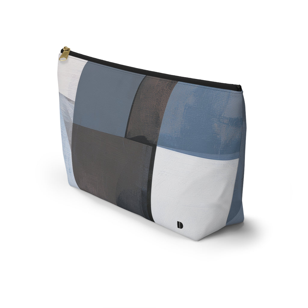 A rectangular zippered pouch with an abstract geometric patchwork pattern in shades of blue, gray, white, and charcoal, featuring a gold zipper pull and a small black logo on the bottom right.