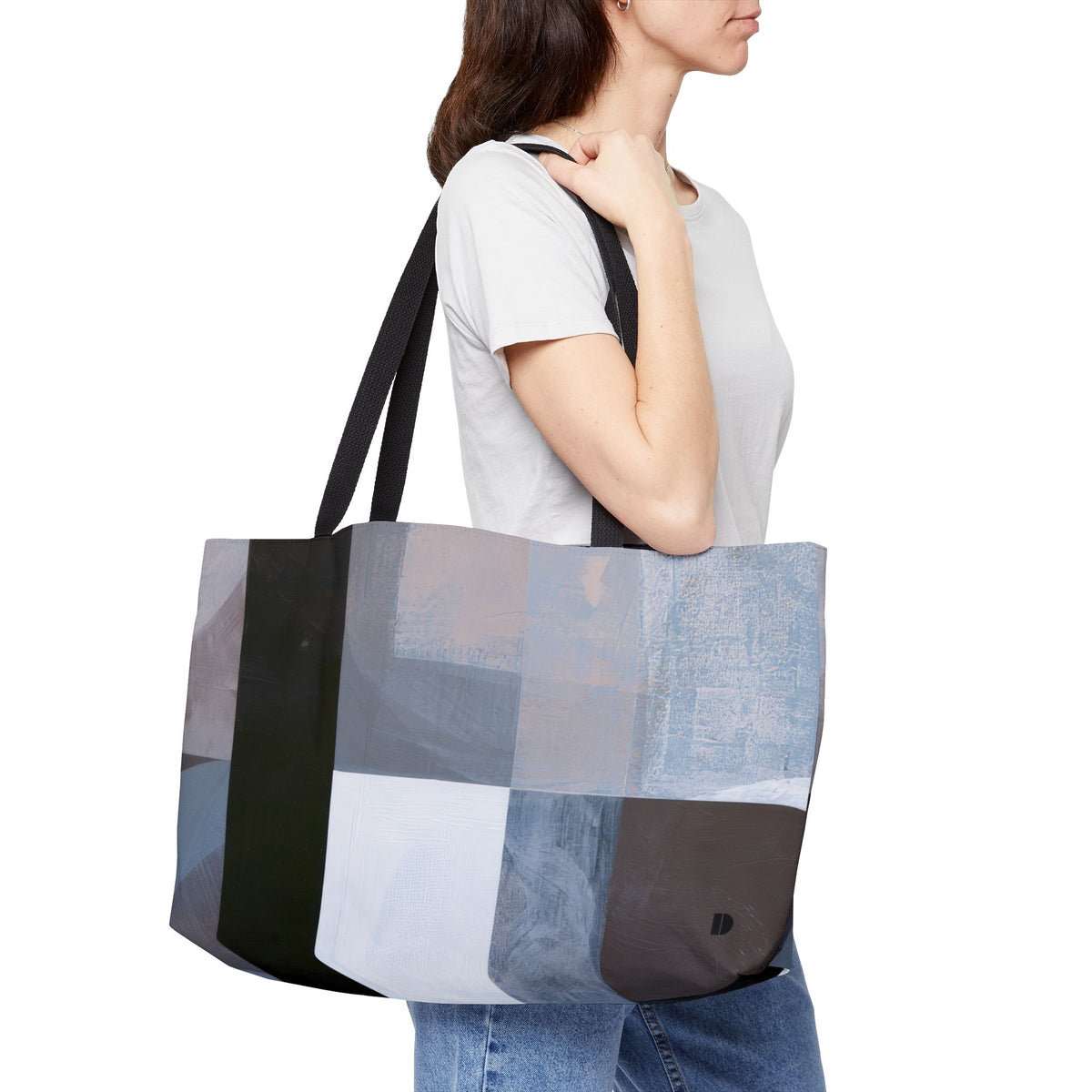 A woman wearing a white t-shirt and blue jeans carries a large patchwork tote bag with black straps. The tote bag features a modern abstract patchwork design in shades of blue, gray, black, and white.
