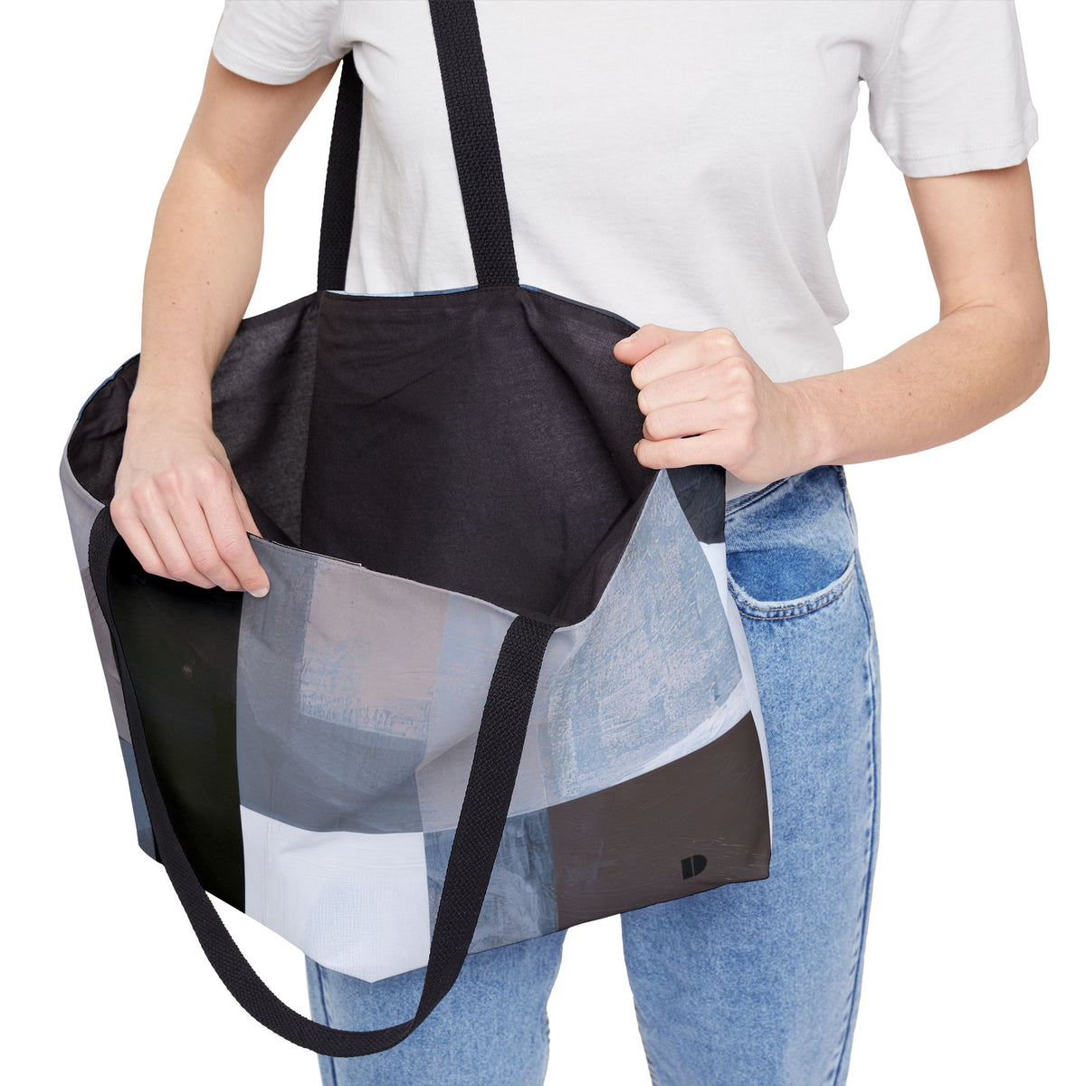 A woman wearing a white t-shirt and blue jeans carries a large patchwork tote bag with black straps and black interior. The tote bag features a modern abstract patchwork design in shades of blue, gray, black, and white.