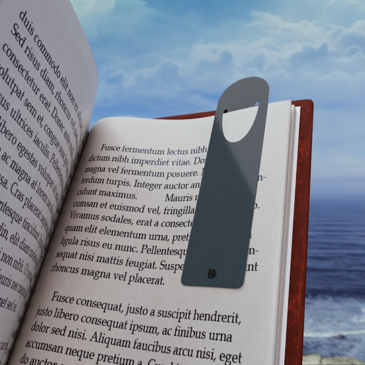 Slate blue metal bookmark shown clipped onto the page of an open book with lorem ipsum text, set against a scenic backdrop of blue ocean waves and sky. Metal bookmark titled 'Slate Blue Metal Bookmark', available in one size.