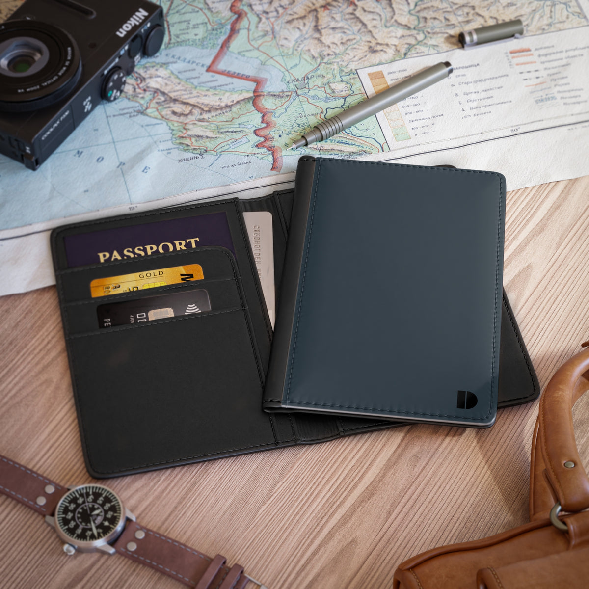 A slate blue faux leather passport holder and card case shown open on a wooden surface, with a passport, cards, camera, map, pen, watch, and leather bag nearby as lifestyle props.