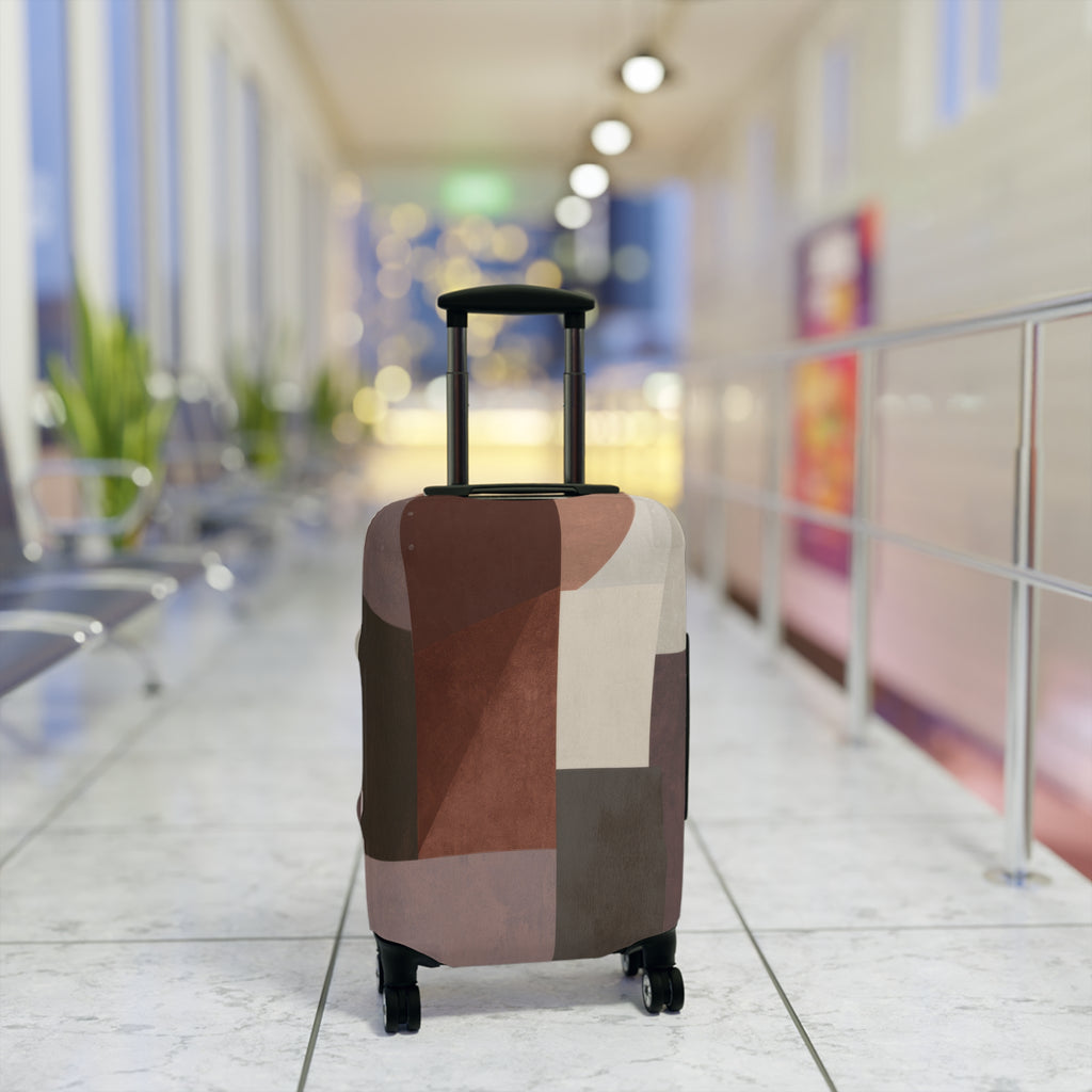 A hard-shell rolling suitcase with an abstract geometric patchwork pattern luggage cover in earthy tones of brown, beige, and gray standing upright in a brightly lit airport terminal.