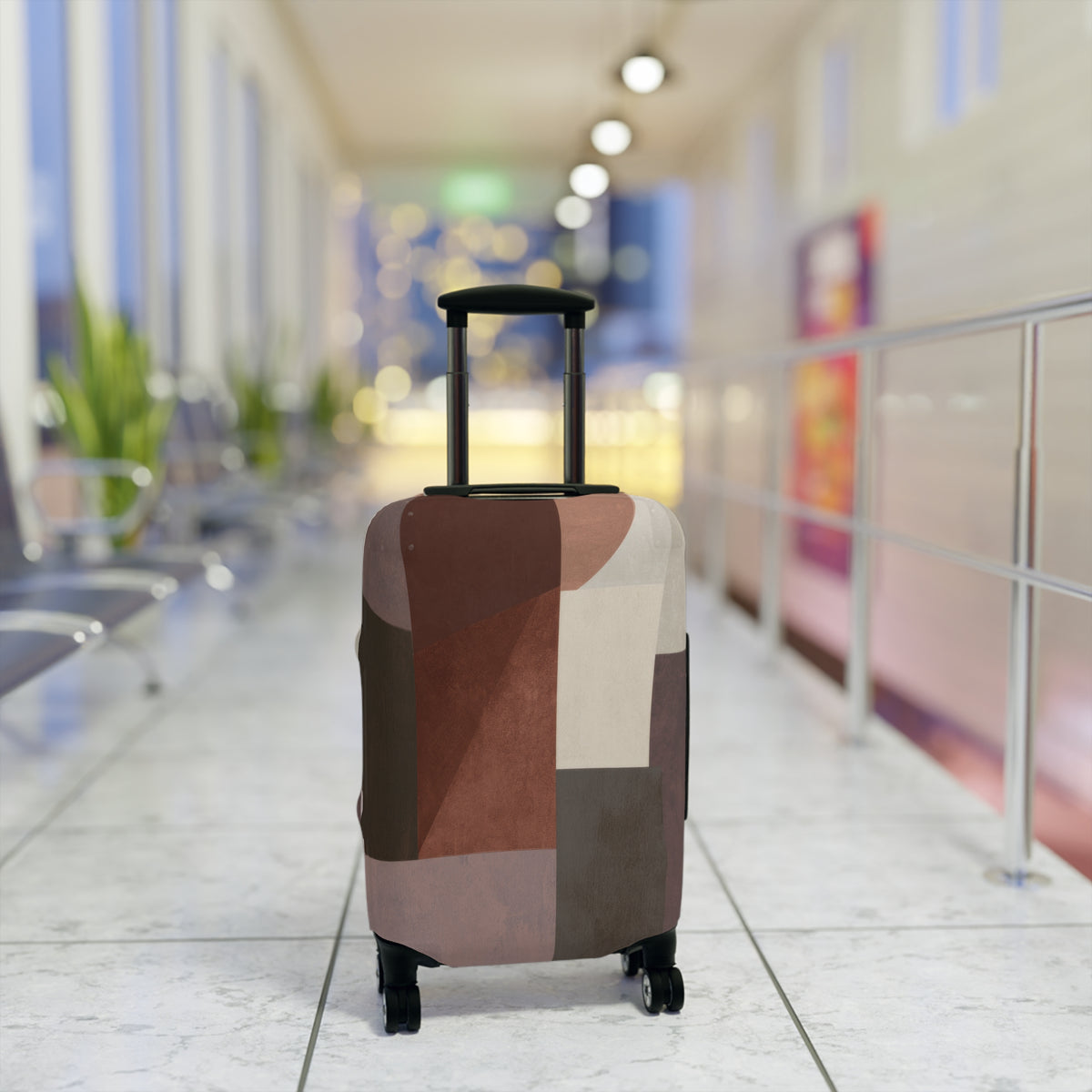 A hard-shell rolling suitcase with an abstract geometric patchwork pattern luggage cover in earthy tones of brown, beige, and gray standing upright in a brightly lit airport terminal.
