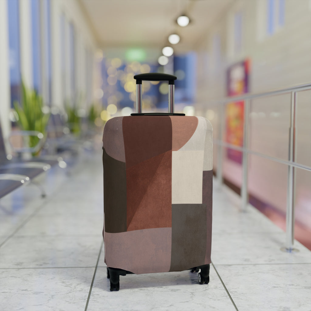 A hard-shell rolling suitcase with an abstract geometric patchwork pattern luggage cover in earthy tones of brown, beige, and gray standing upright in a brightly lit airport terminal.
