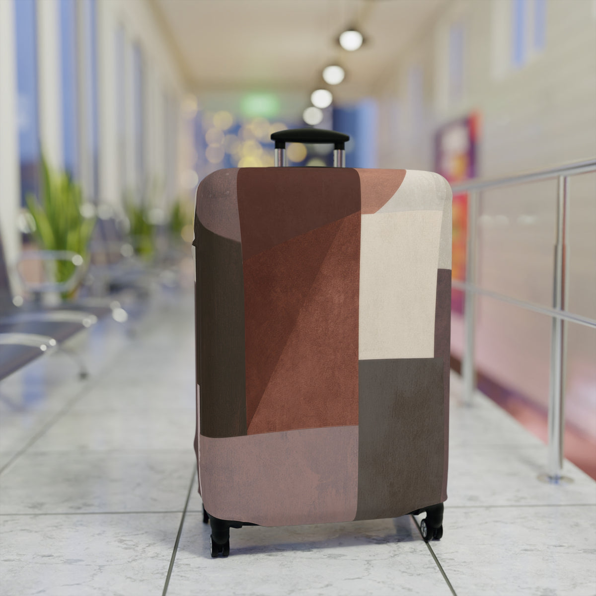A hard-shell rolling suitcase with an abstract geometric patchwork pattern luggage cover in earthy tones of brown, beige, and gray standing upright in a brightly lit airport terminal.