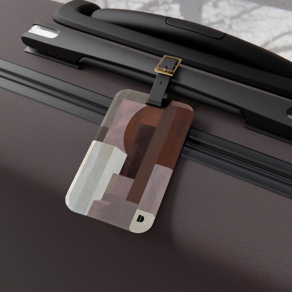 A rectangular luggage tag with rounded corners and a geometric abstract pattern in shades of brown, beige, and gray, attached to a black strap on a dark brown textured suitcase.