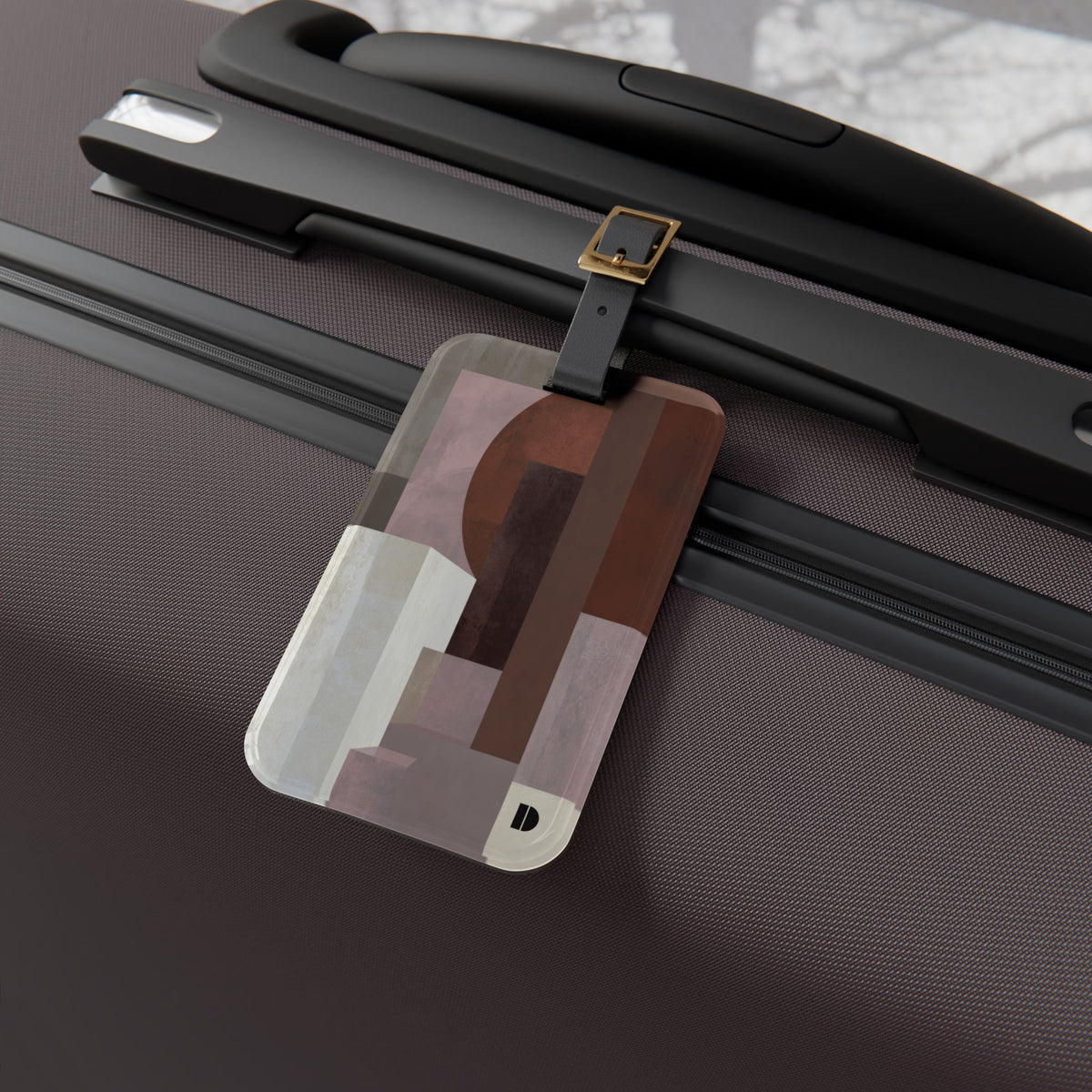 A rectangular luggage tag with rounded corners and a geometric abstract pattern in shades of brown, beige, and gray, attached to a black strap on a dark brown textured suitcase.