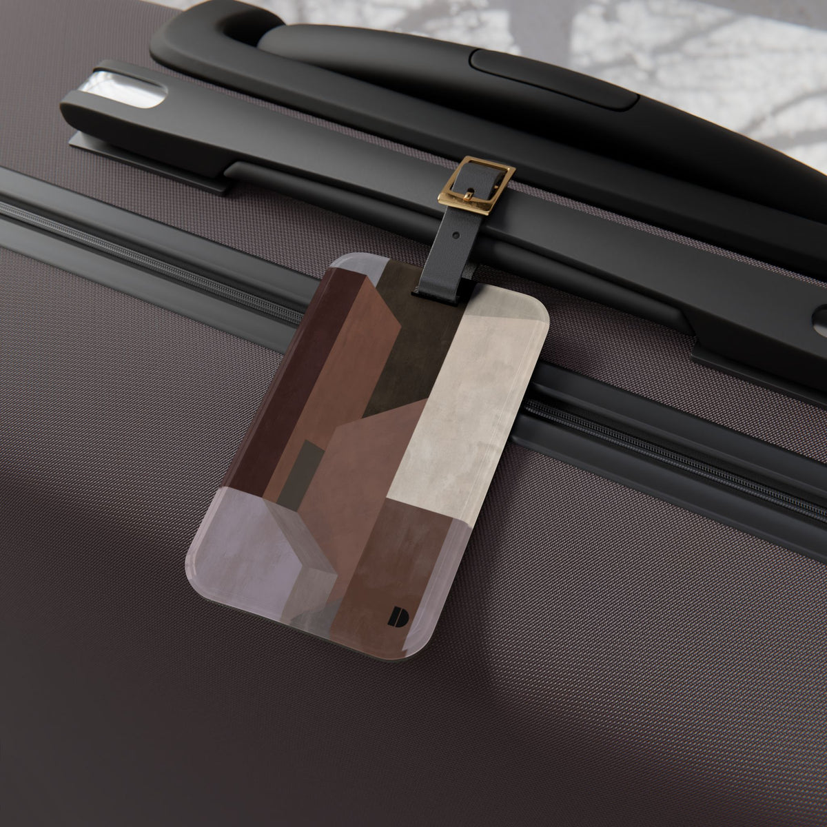A rectangular luggage tag with rounded corners hangs from a black strap on the handle of a textured dark brown suitcase. The tag features an abstract geometric pattern in shades of brown, beige, and gray, with a small black logo at the bottom right.