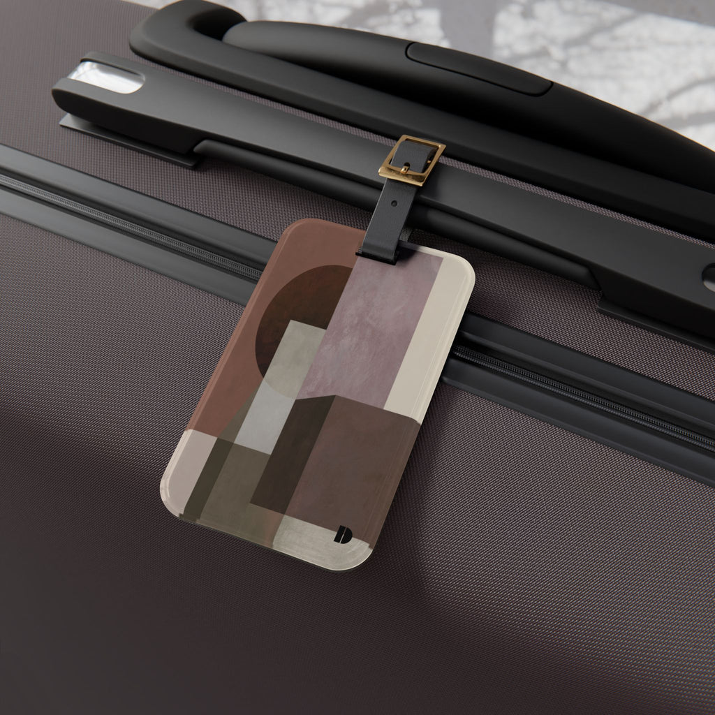 A rectangular luggage tag with an abstract geometric pattern in brown, beige, and muted pink, attached to a black strap on a dark textured suitcase.