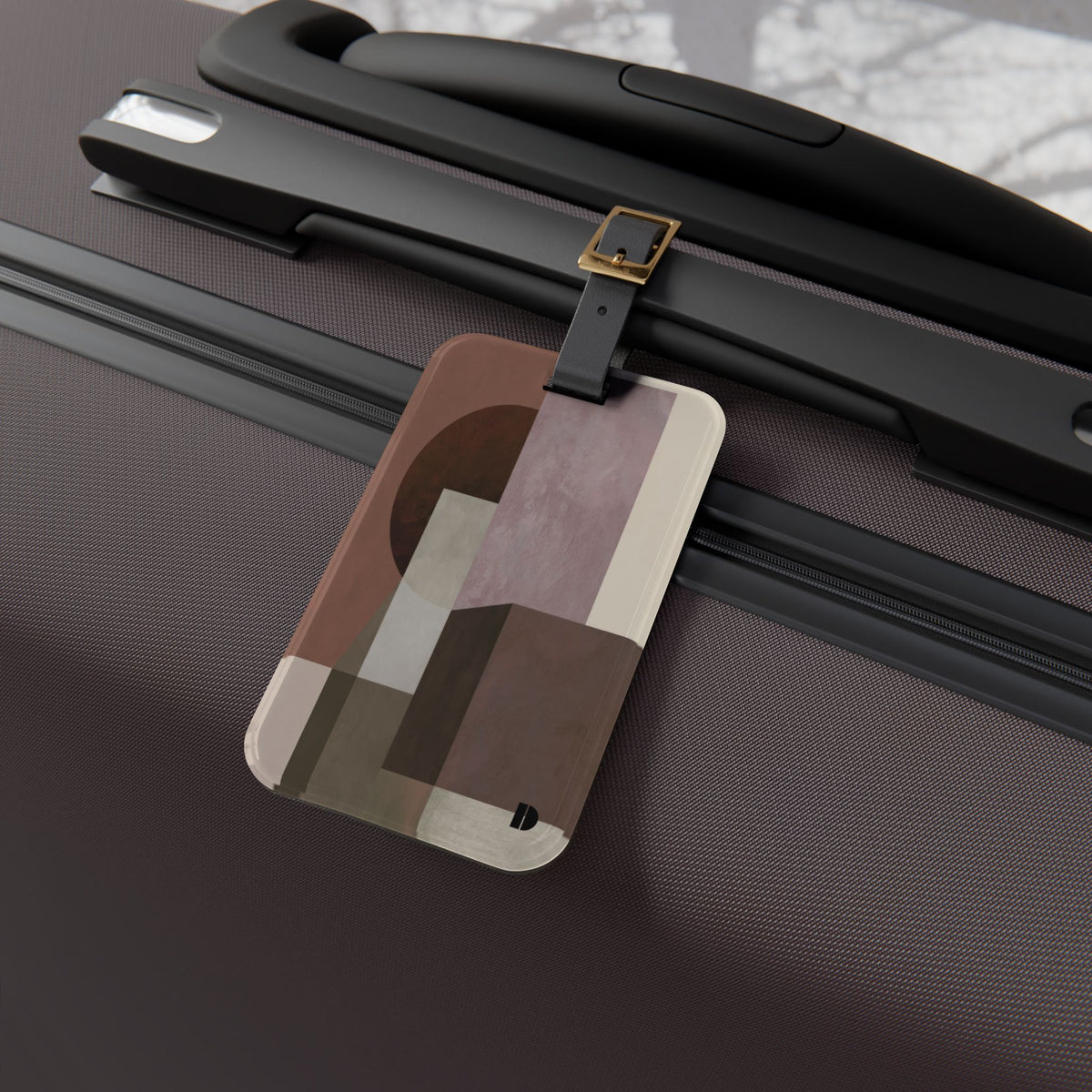 A rectangular luggage tag with an abstract geometric pattern in brown, beige, and muted pink, attached to a black strap on a dark textured suitcase.