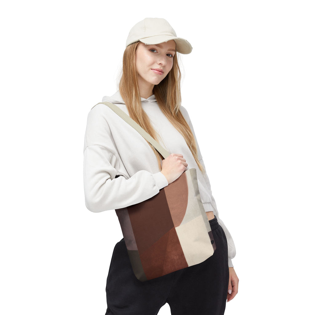 A woman wearing a beige cap, white hoodie, and black sweatpants carries a large canvas tote bag with a geometric patchwork pattern in shades of brown, beige, and gray. The photo is shot against a solid white background.