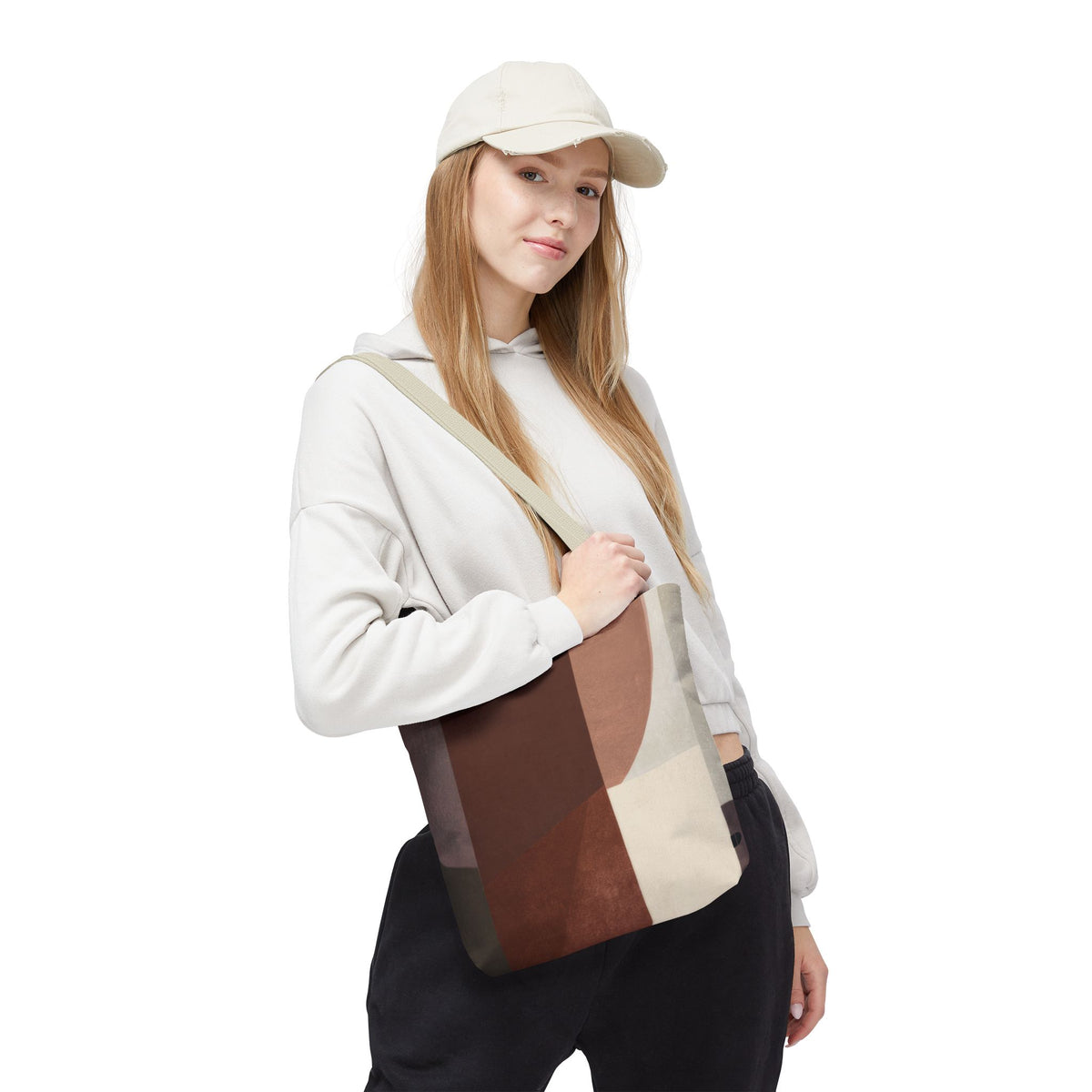 A woman wearing a beige cap, white hoodie, and black sweatpants carries a large canvas tote bag with a geometric patchwork pattern in shades of brown, beige, and gray. The photo is shot against a solid white background.