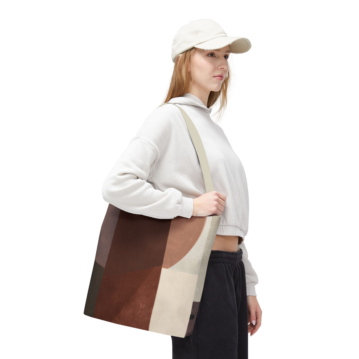 A woman wearing a white cap, white sweatshirt, and black sweatpants carries a large canvas tote bag with a geometric patchwork pattern in brown, beige, and gray. The photo is shot against a seamless white background.