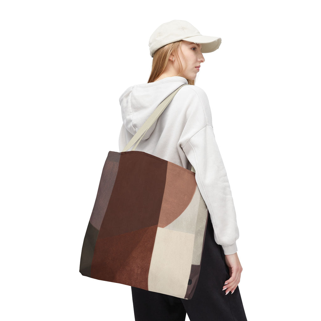 A woman wearing a white cap, white sweatshirt, and black sweatpants carries a large canvas tote bag with a geometric patchwork pattern in brown, beige, and gray. The photo is shot against a seamless white background.