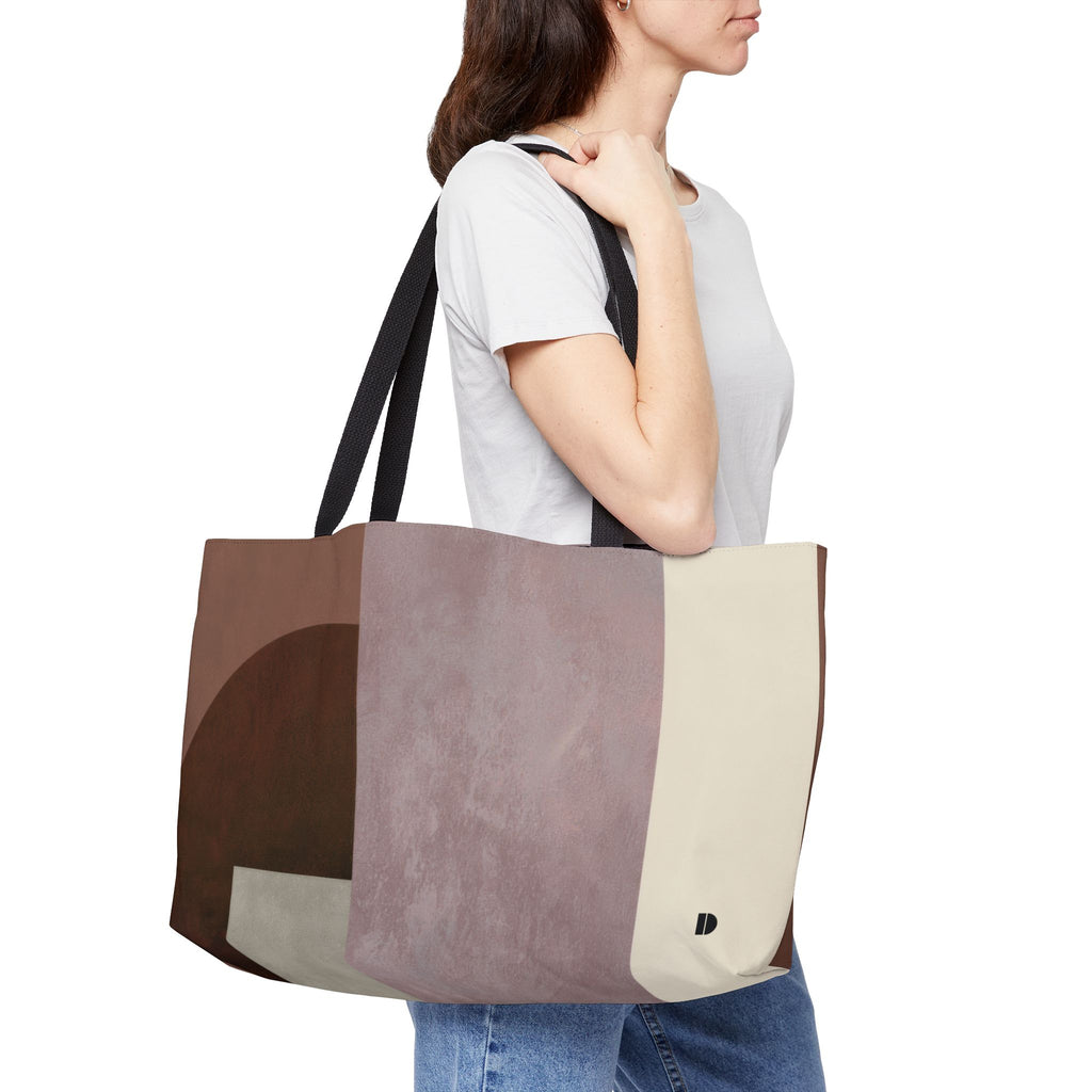A woman wearing a white t-shirt and blue jeans carries a large, structured tote bag with a geometric color-blocked pattern in brown, dusty mauve, and cream. The bag has black shoulder straps and a small black monogram logo on the lower right corner.