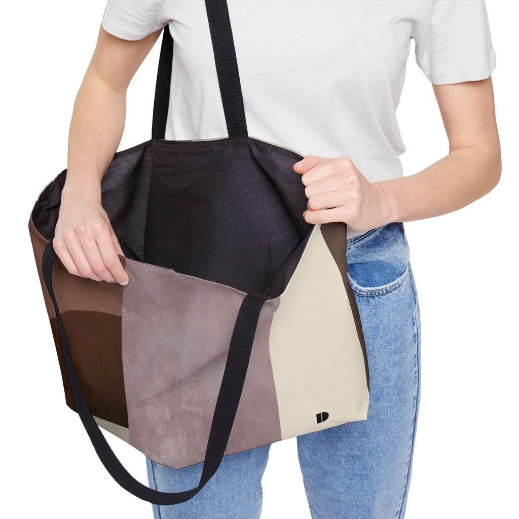 A person wearing a white t-shirt and blue jeans holds open a large patchwork tote bag with black shoulder straps and black interior. The bag features color-blocked panels in black, brown, taupe, beige, and light gray, shown against a plain white background.